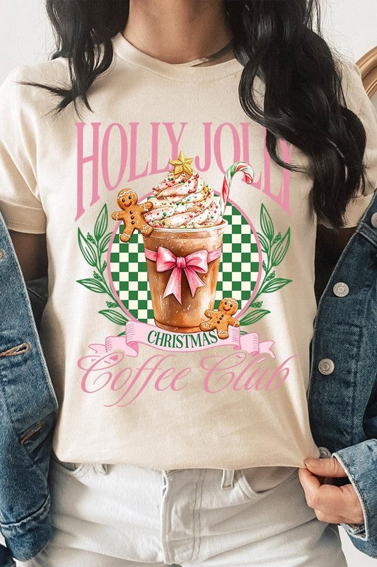 A woman with pink hair is wearing the "Holly Jolly Christmas Coffee Club Graphic Tee," adorned with a charming design of a festive coffee cup and gingerbread men. The Christmas decorations in the background enhance the jolly atmosphere of this unisex crew neck shirt.