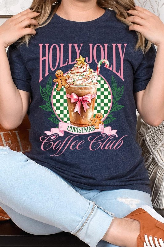 A woman with pink hair is wearing the "Holly Jolly Christmas Coffee Club Graphic Tee," adorned with a charming design of a festive coffee cup and gingerbread men. The Christmas decorations in the background enhance the jolly atmosphere of this unisex crew neck shirt.