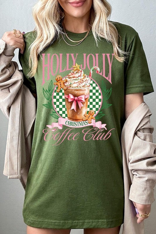 A woman with pink hair is wearing the "Holly Jolly Christmas Coffee Club Graphic Tee," adorned with a charming design of a festive coffee cup and gingerbread men. The Christmas decorations in the background enhance the jolly atmosphere of this unisex crew neck shirt.