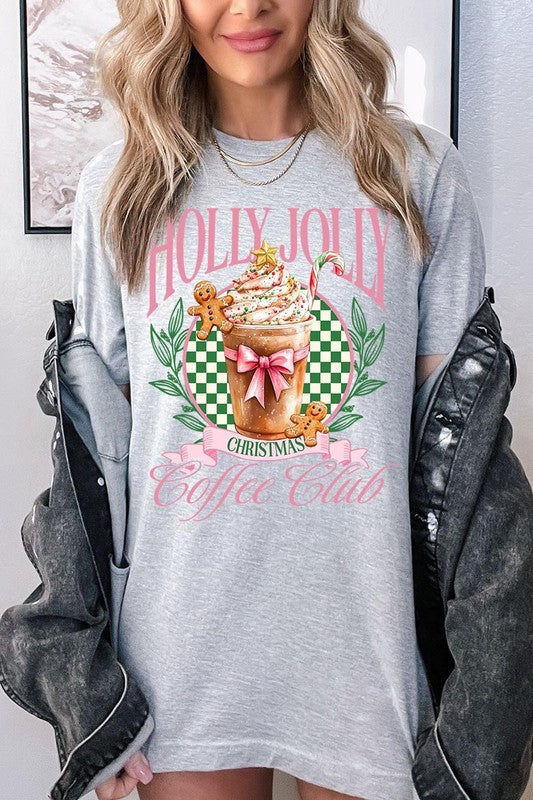 A woman with pink hair is wearing the "Holly Jolly Christmas Coffee Club Graphic Tee," adorned with a charming design of a festive coffee cup and gingerbread men. The Christmas decorations in the background enhance the jolly atmosphere of this unisex crew neck shirt.