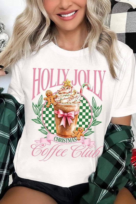 A woman with pink hair is wearing the "Holly Jolly Christmas Coffee Club Graphic Tee," adorned with a charming design of a festive coffee cup and gingerbread men. The Christmas decorations in the background enhance the jolly atmosphere of this unisex crew neck shirt.