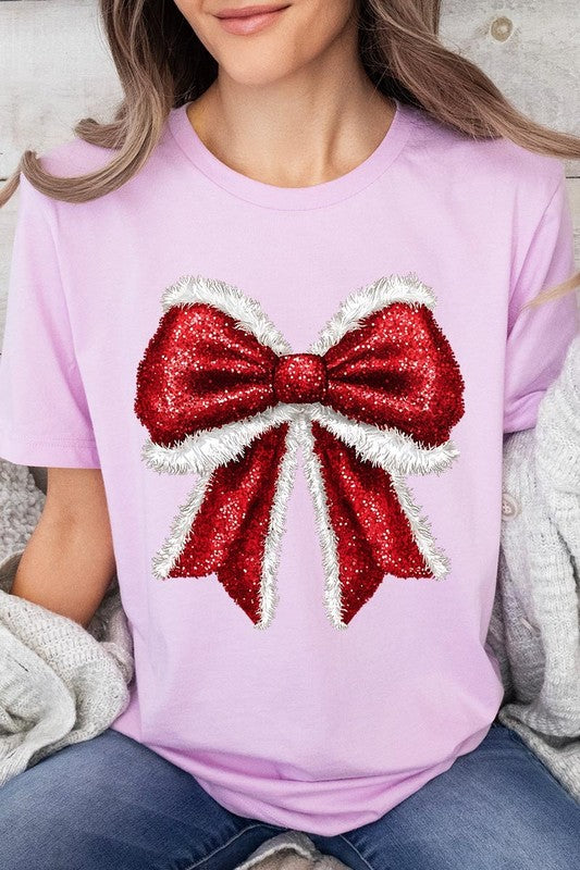 A woman wears a cheerful Christmas Santa Bow Graphic Tee, showcasing a prominent glittery bow design on the front. Complementing her attire, she has accessorized with a gleaming silver chain necklace and wrapped herself in a snug white blanket over one shoulder. Perfect for Family Group Uniforms, this ensemble exudes festive spirit.