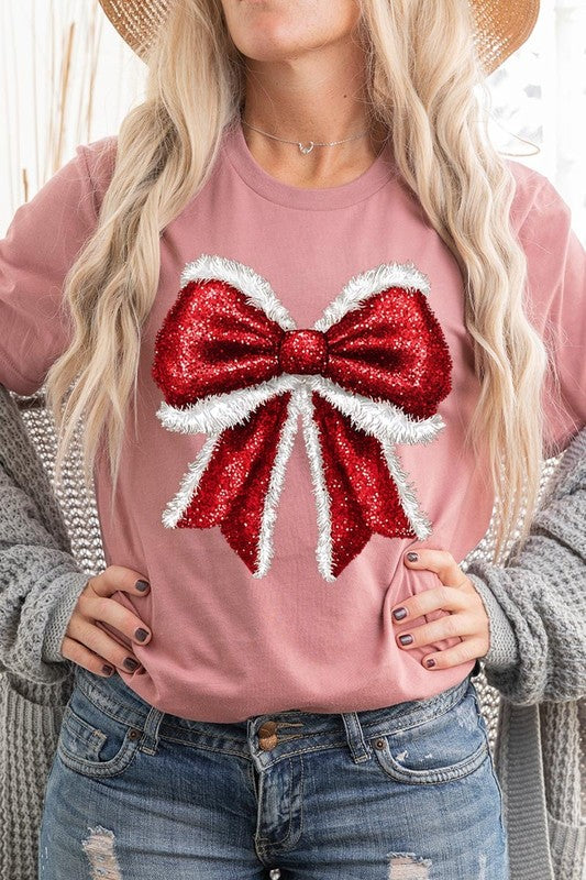 A woman wears a cheerful Christmas Santa Bow Graphic Tee, showcasing a prominent glittery bow design on the front. Complementing her attire, she has accessorized with a gleaming silver chain necklace and wrapped herself in a snug white blanket over one shoulder. Perfect for Family Group Uniforms, this ensemble exudes festive spirit.