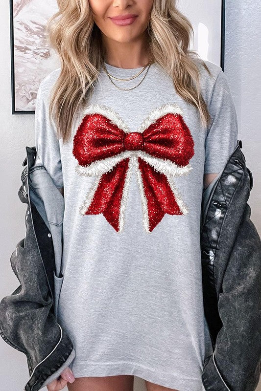 A woman wears a cheerful Christmas Santa Bow Graphic Tee, showcasing a prominent glittery bow design on the front. Complementing her attire, she has accessorized with a gleaming silver chain necklace and wrapped herself in a snug white blanket over one shoulder. Perfect for Family Group Uniforms, this ensemble exudes festive spirit.