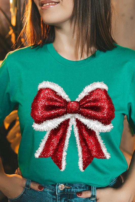 A woman wears a cheerful Christmas Santa Bow Graphic Tee, showcasing a prominent glittery bow design on the front. Complementing her attire, she has accessorized with a gleaming silver chain necklace and wrapped herself in a snug white blanket over one shoulder. Perfect for Family Group Uniforms, this ensemble exudes festive spirit.