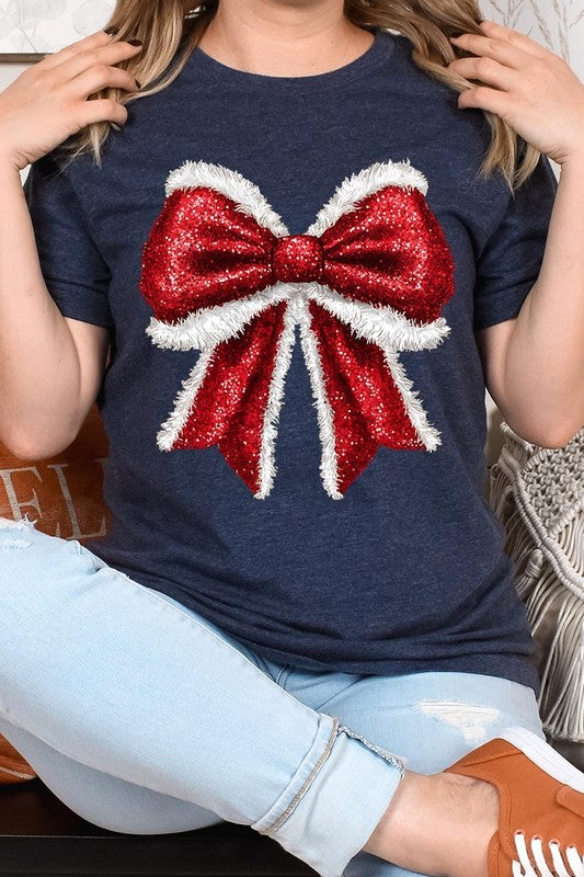 A woman wears a cheerful Christmas Santa Bow Graphic Tee, showcasing a prominent glittery bow design on the front. Complementing her attire, she has accessorized with a gleaming silver chain necklace and wrapped herself in a snug white blanket over one shoulder. Perfect for Family Group Uniforms, this ensemble exudes festive spirit.