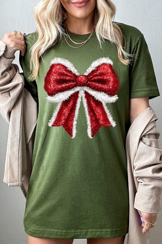 A woman wears a cheerful Christmas Santa Bow Graphic Tee, showcasing a prominent glittery bow design on the front. Complementing her attire, she has accessorized with a gleaming silver chain necklace and wrapped herself in a snug white blanket over one shoulder. Perfect for Family Group Uniforms, this ensemble exudes festive spirit.