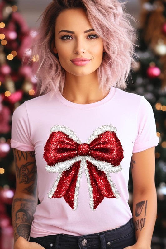 A woman wears a cheerful Christmas Santa Bow Graphic Tee, showcasing a prominent glittery bow design on the front. Complementing her attire, she has accessorized with a gleaming silver chain necklace and wrapped herself in a snug white blanket over one shoulder. Perfect for Family Group Uniforms, this ensemble exudes festive spirit.