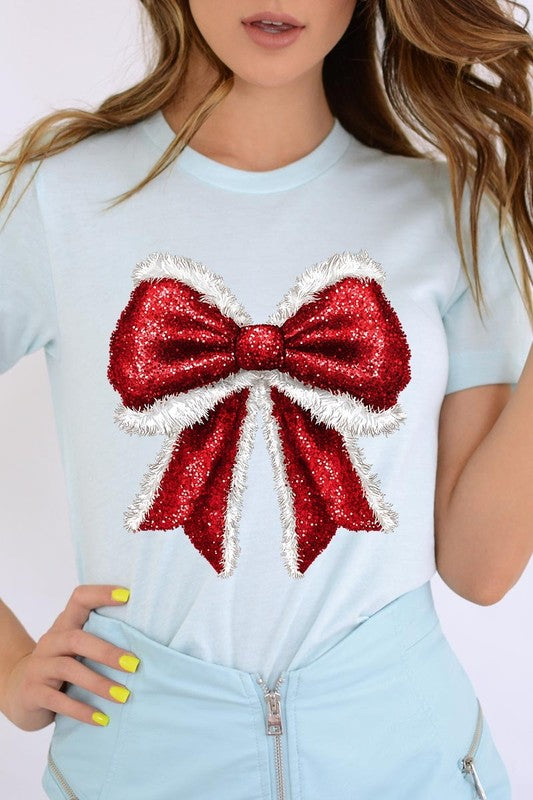 A woman wears a cheerful Christmas Santa Bow Graphic Tee, showcasing a prominent glittery bow design on the front. Complementing her attire, she has accessorized with a gleaming silver chain necklace and wrapped herself in a snug white blanket over one shoulder. Perfect for Family Group Uniforms, this ensemble exudes festive spirit.