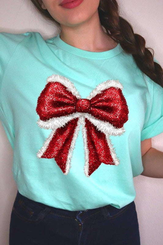 A woman wears a cheerful Christmas Santa Bow Graphic Tee, showcasing a prominent glittery bow design on the front. Complementing her attire, she has accessorized with a gleaming silver chain necklace and wrapped herself in a snug white blanket over one shoulder. Perfect for Family Group Uniforms, this ensemble exudes festive spirit.