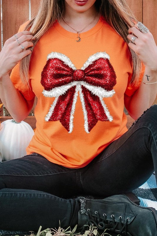 A woman wears a cheerful Christmas Santa Bow Graphic Tee, showcasing a prominent glittery bow design on the front. Complementing her attire, she has accessorized with a gleaming silver chain necklace and wrapped herself in a snug white blanket over one shoulder. Perfect for Family Group Uniforms, this ensemble exudes festive spirit.