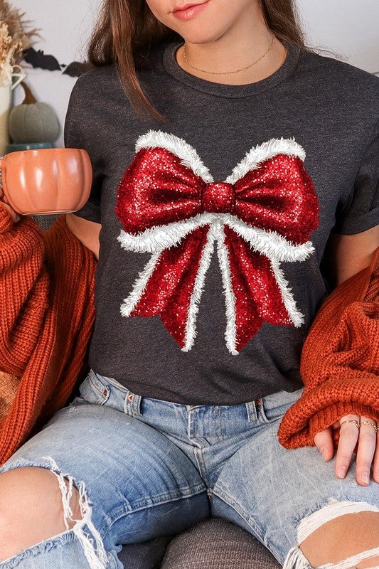 A woman wears a cheerful Christmas Santa Bow Graphic Tee, showcasing a prominent glittery bow design on the front. Complementing her attire, she has accessorized with a gleaming silver chain necklace and wrapped herself in a snug white blanket over one shoulder. Perfect for Family Group Uniforms, this ensemble exudes festive spirit.