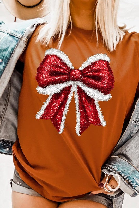 A woman wears a cheerful Christmas Santa Bow Graphic Tee, showcasing a prominent glittery bow design on the front. Complementing her attire, she has accessorized with a gleaming silver chain necklace and wrapped herself in a snug white blanket over one shoulder. Perfect for Family Group Uniforms, this ensemble exudes festive spirit.