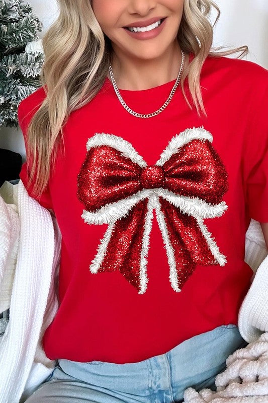 A woman wears a cheerful Christmas Santa Bow Graphic Tee, showcasing a prominent glittery bow design on the front. Complementing her attire, she has accessorized with a gleaming silver chain necklace and wrapped herself in a snug white blanket over one shoulder. Perfect for Family Group Uniforms, this ensemble exudes festive spirit.