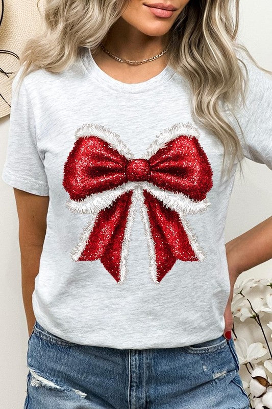 A woman wears a cheerful Christmas Santa Bow Graphic Tee, showcasing a prominent glittery bow design on the front. Complementing her attire, she has accessorized with a gleaming silver chain necklace and wrapped herself in a snug white blanket over one shoulder. Perfect for Family Group Uniforms, this ensemble exudes festive spirit.