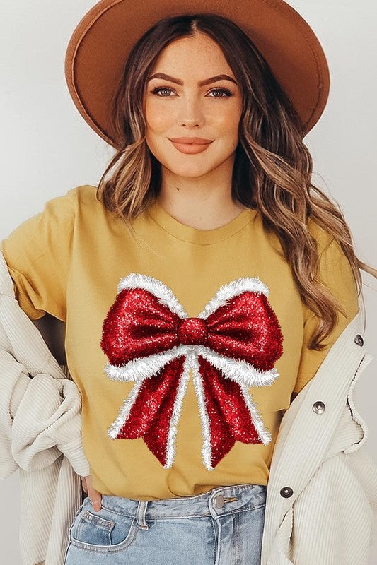 A woman wears a cheerful Christmas Santa Bow Graphic Tee, showcasing a prominent glittery bow design on the front. Complementing her attire, she has accessorized with a gleaming silver chain necklace and wrapped herself in a snug white blanket over one shoulder. Perfect for Family Group Uniforms, this ensemble exudes festive spirit.