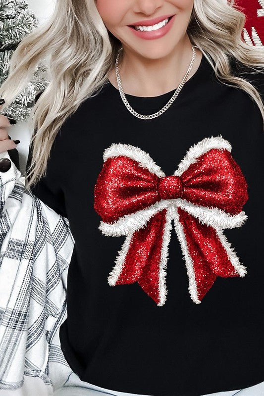 A woman wears a cheerful Christmas Santa Bow Graphic Tee, showcasing a prominent glittery bow design on the front. Complementing her attire, she has accessorized with a gleaming silver chain necklace and wrapped herself in a snug white blanket over one shoulder. Perfect for Family Group Uniforms, this ensemble exudes festive spirit.