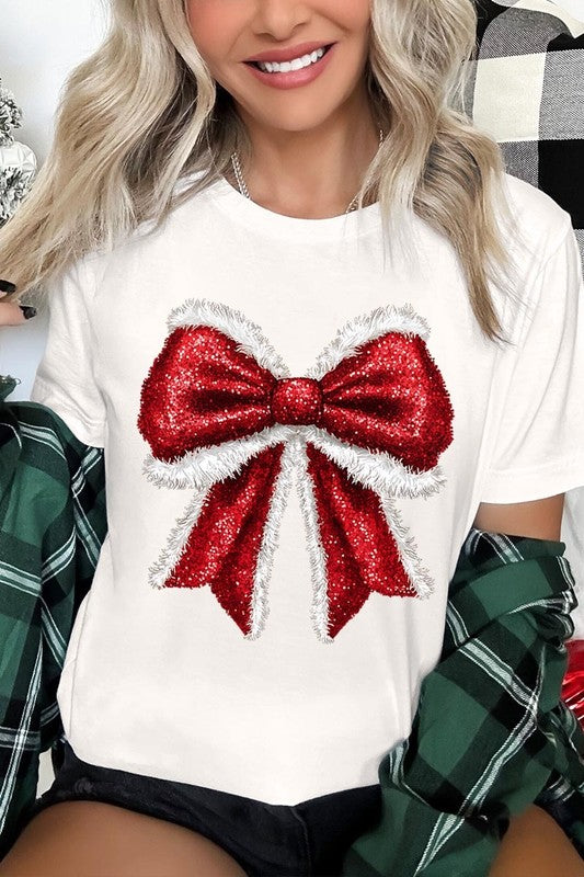 A woman wears a cheerful Christmas Santa Bow Graphic Tee, showcasing a prominent glittery bow design on the front. Complementing her attire, she has accessorized with a gleaming silver chain necklace and wrapped herself in a snug white blanket over one shoulder. Perfect for Family Group Uniforms, this ensemble exudes festive spirit.