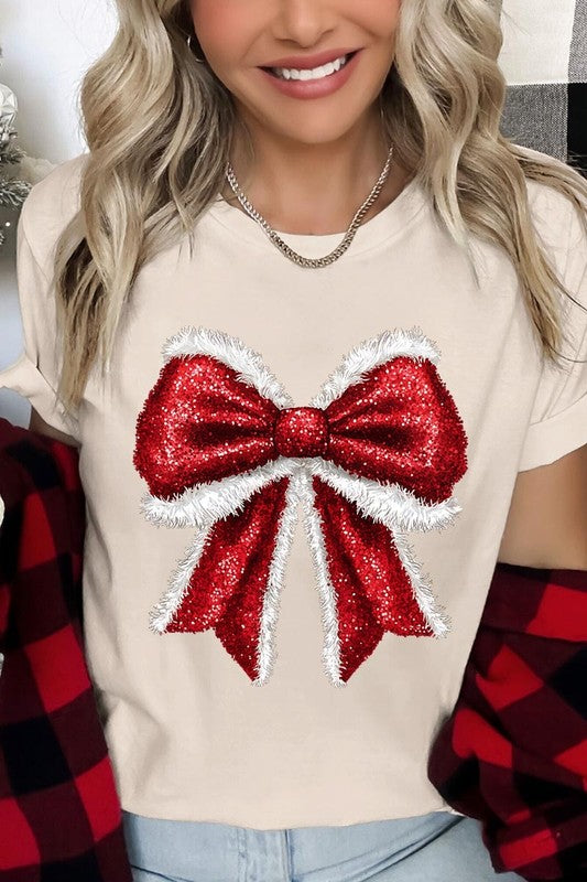 A woman wears a cheerful Christmas Santa Bow Graphic Tee, showcasing a prominent glittery bow design on the front. Complementing her attire, she has accessorized with a gleaming silver chain necklace and wrapped herself in a snug white blanket over one shoulder. Perfect for Family Group Uniforms, this ensemble exudes festive spirit.