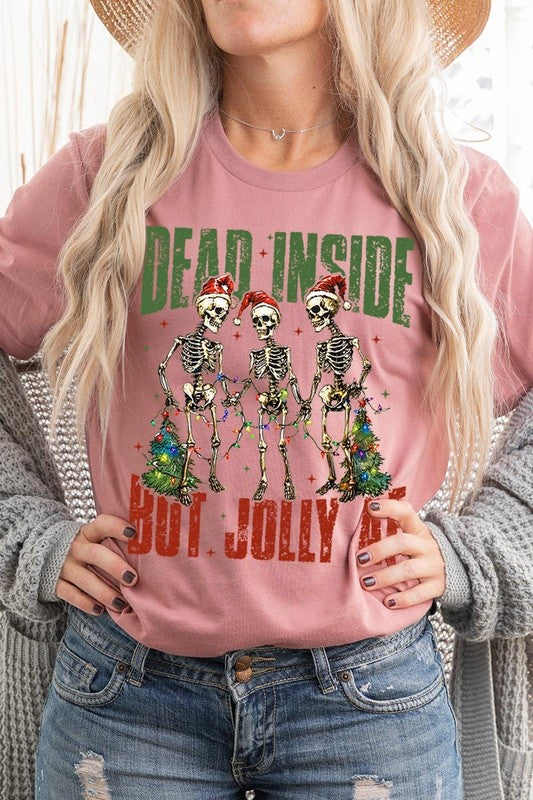 A person wearing the Dead Inside But Jolly AF Skeleton Graphic Tee, which features a beige color and a graphic of three festive skeletons holding string lights and Christmas trees, adorned with the text "Dead Inside But Jolly AF." This unisex crew neck tee offers a comfortable fit, making it perfect for adding some humor to your holiday wardrobe.