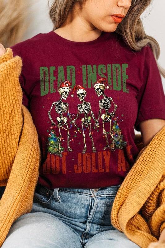 A person wearing the Dead Inside But Jolly AF Skeleton Graphic Tee, which features a beige color and a graphic of three festive skeletons holding string lights and Christmas trees, adorned with the text "Dead Inside But Jolly AF." This unisex crew neck tee offers a comfortable fit, making it perfect for adding some humor to your holiday wardrobe.