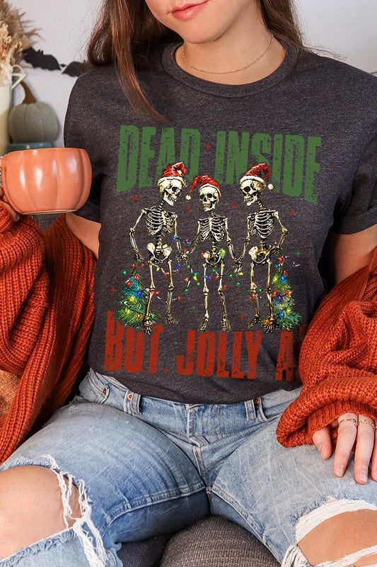 A person wearing the Dead Inside But Jolly AF Skeleton Graphic Tee, which features a beige color and a graphic of three festive skeletons holding string lights and Christmas trees, adorned with the text "Dead Inside But Jolly AF." This unisex crew neck tee offers a comfortable fit, making it perfect for adding some humor to your holiday wardrobe.