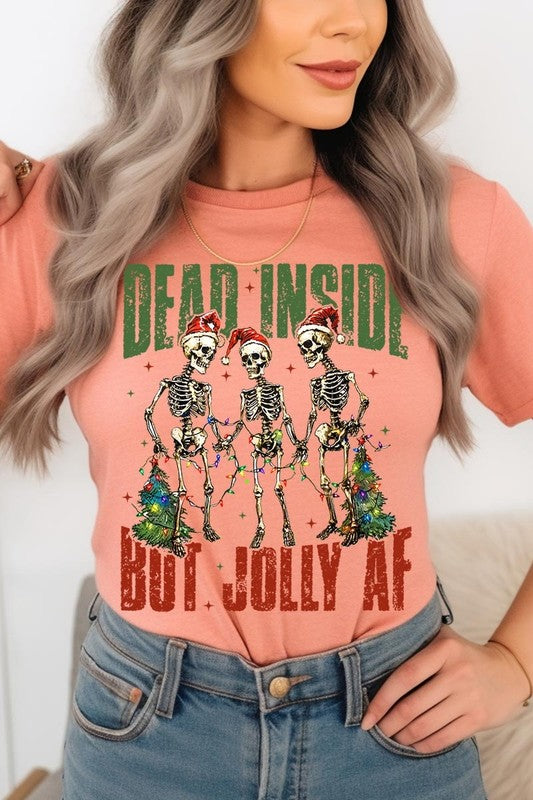 A person wearing the Dead Inside But Jolly AF Skeleton Graphic Tee, which features a beige color and a graphic of three festive skeletons holding string lights and Christmas trees, adorned with the text "Dead Inside But Jolly AF." This unisex crew neck tee offers a comfortable fit, making it perfect for adding some humor to your holiday wardrobe.