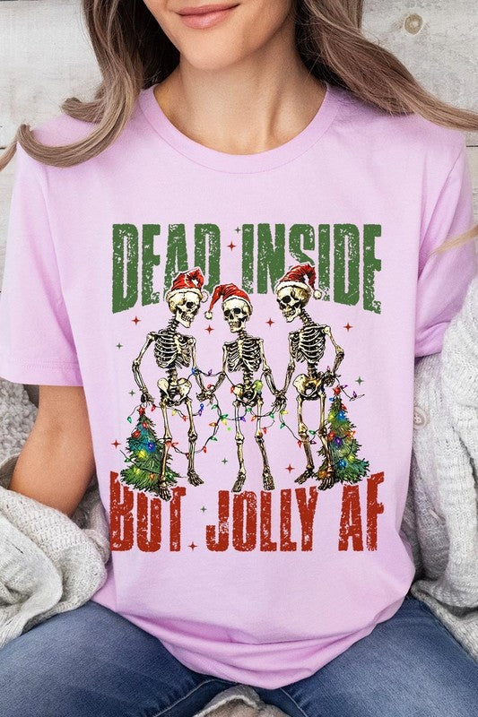 A person wearing the Dead Inside But Jolly AF Skeleton Graphic Tee, which features a beige color and a graphic of three festive skeletons holding string lights and Christmas trees, adorned with the text "Dead Inside But Jolly AF." This unisex crew neck tee offers a comfortable fit, making it perfect for adding some humor to your holiday wardrobe.