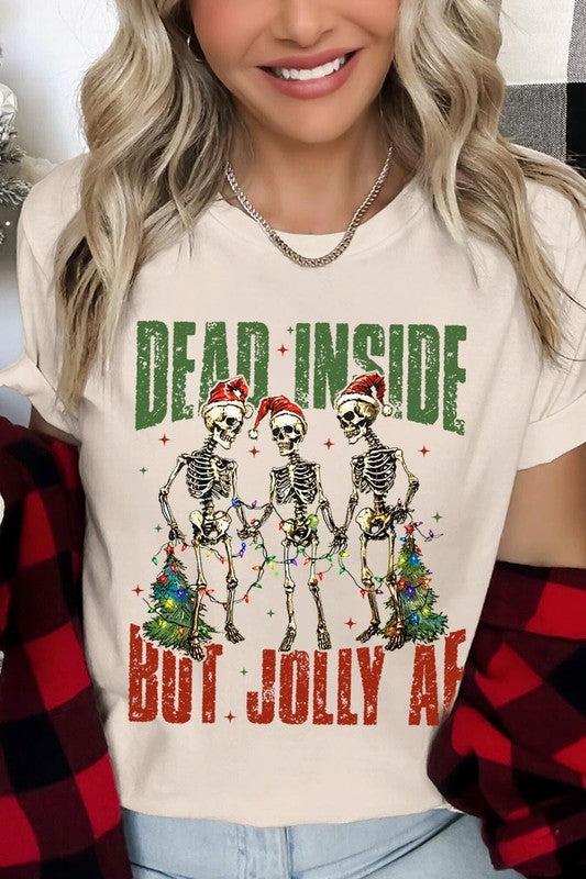 A person wearing the Dead Inside But Jolly AF Skeleton Graphic Tee, which features a beige color and a graphic of three festive skeletons holding string lights and Christmas trees, adorned with the text "Dead Inside But Jolly AF." This unisex crew neck tee offers a comfortable fit, making it perfect for adding some humor to your holiday wardrobe.