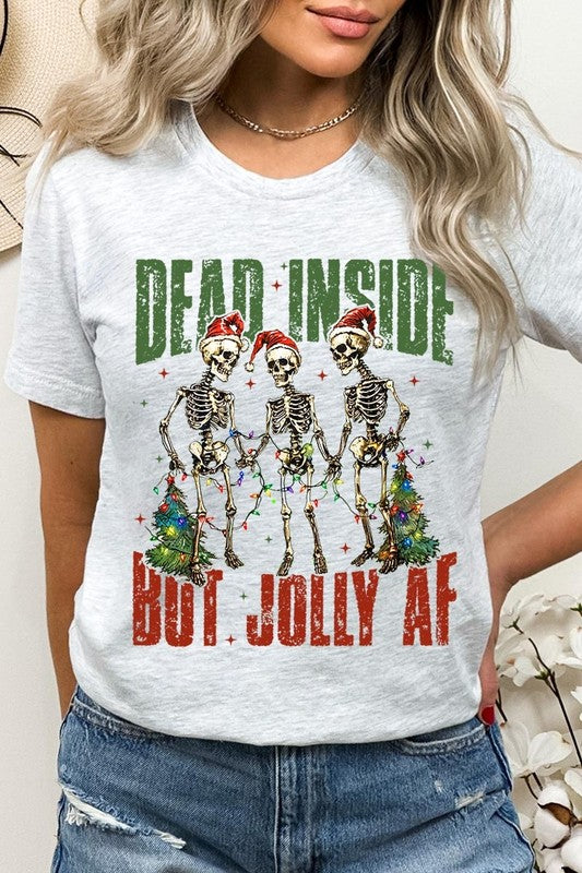 A person wearing the Dead Inside But Jolly AF Skeleton Graphic Tee, which features a beige color and a graphic of three festive skeletons holding string lights and Christmas trees, adorned with the text "Dead Inside But Jolly AF." This unisex crew neck tee offers a comfortable fit, making it perfect for adding some humor to your holiday wardrobe.