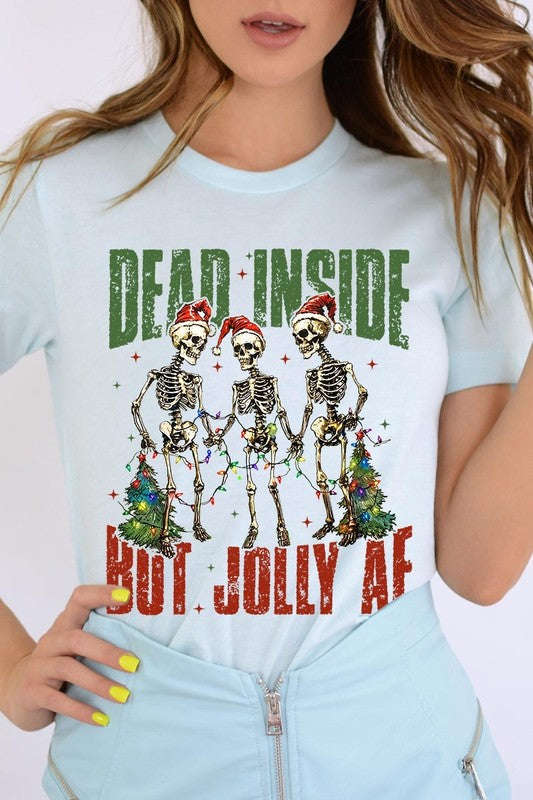 A person wearing the Dead Inside But Jolly AF Skeleton Graphic Tee, which features a beige color and a graphic of three festive skeletons holding string lights and Christmas trees, adorned with the text "Dead Inside But Jolly AF." This unisex crew neck tee offers a comfortable fit, making it perfect for adding some humor to your holiday wardrobe.