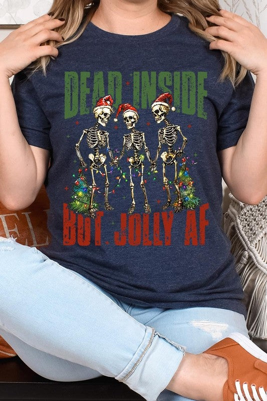 A person wearing the Dead Inside But Jolly AF Skeleton Graphic Tee, which features a beige color and a graphic of three festive skeletons holding string lights and Christmas trees, adorned with the text "Dead Inside But Jolly AF." This unisex crew neck tee offers a comfortable fit, making it perfect for adding some humor to your holiday wardrobe.