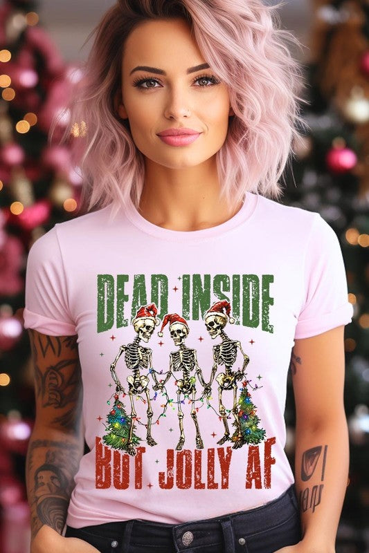 A person wearing the Dead Inside But Jolly AF Skeleton Graphic Tee, which features a beige color and a graphic of three festive skeletons holding string lights and Christmas trees, adorned with the text "Dead Inside But Jolly AF." This unisex crew neck tee offers a comfortable fit, making it perfect for adding some humor to your holiday wardrobe.