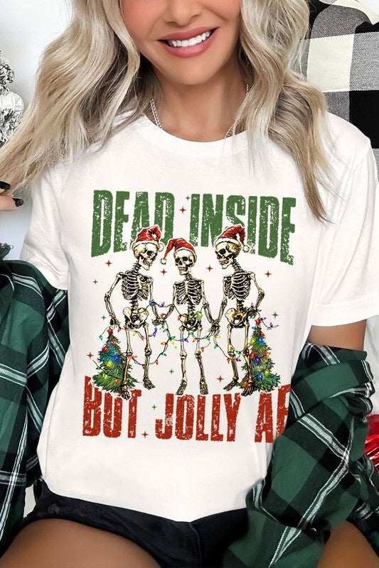 A person wearing the Dead Inside But Jolly AF Skeleton Graphic Tee, which features a beige color and a graphic of three festive skeletons holding string lights and Christmas trees, adorned with the text "Dead Inside But Jolly AF." This unisex crew neck tee offers a comfortable fit, making it perfect for adding some humor to your holiday wardrobe.