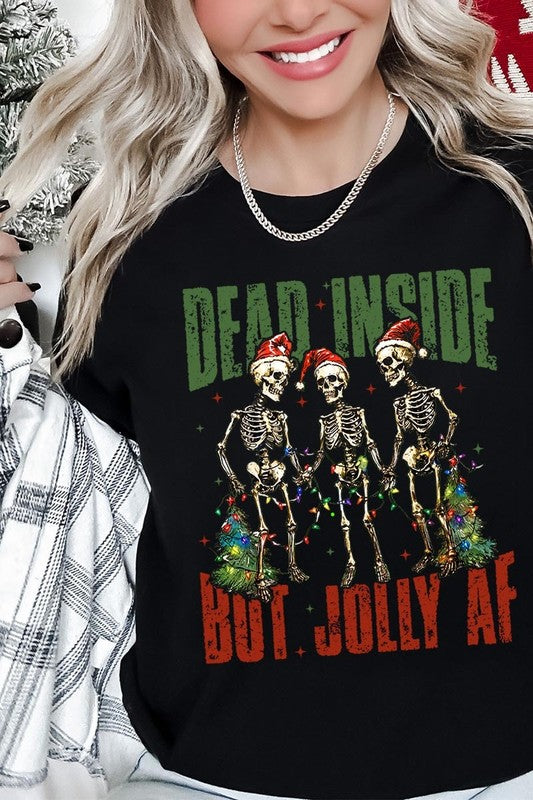 A person wearing the Dead Inside But Jolly AF Skeleton Graphic Tee, which features a beige color and a graphic of three festive skeletons holding string lights and Christmas trees, adorned with the text "Dead Inside But Jolly AF." This unisex crew neck tee offers a comfortable fit, making it perfect for adding some humor to your holiday wardrobe.