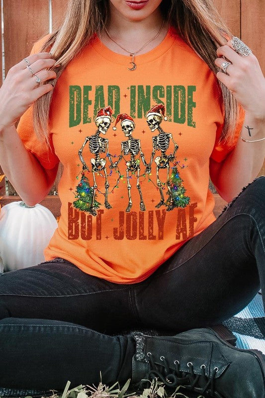 A person wearing the Dead Inside But Jolly AF Skeleton Graphic Tee, which features a beige color and a graphic of three festive skeletons holding string lights and Christmas trees, adorned with the text "Dead Inside But Jolly AF." This unisex crew neck tee offers a comfortable fit, making it perfect for adding some humor to your holiday wardrobe.