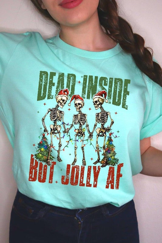 A person wearing the Dead Inside But Jolly AF Skeleton Graphic Tee, which features a beige color and a graphic of three festive skeletons holding string lights and Christmas trees, adorned with the text "Dead Inside But Jolly AF." This unisex crew neck tee offers a comfortable fit, making it perfect for adding some humor to your holiday wardrobe.