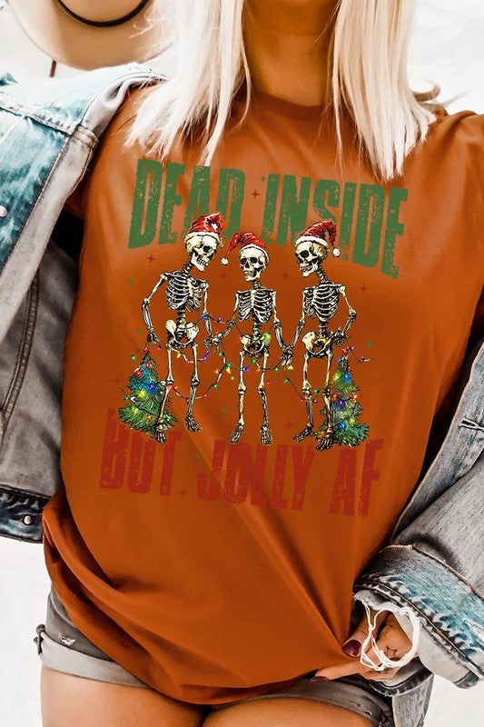 A person wearing the Dead Inside But Jolly AF Skeleton Graphic Tee, which features a beige color and a graphic of three festive skeletons holding string lights and Christmas trees, adorned with the text "Dead Inside But Jolly AF." This unisex crew neck tee offers a comfortable fit, making it perfect for adding some humor to your holiday wardrobe.