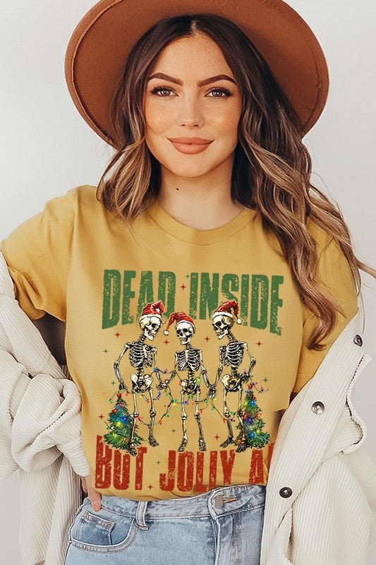 A person wearing the Dead Inside But Jolly AF Skeleton Graphic Tee, which features a beige color and a graphic of three festive skeletons holding string lights and Christmas trees, adorned with the text "Dead Inside But Jolly AF." This unisex crew neck tee offers a comfortable fit, making it perfect for adding some humor to your holiday wardrobe.