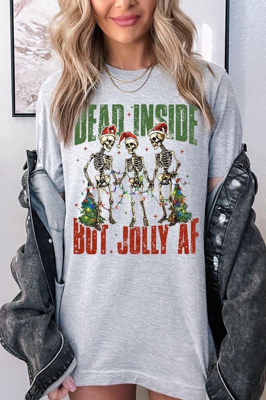 A person wearing the Dead Inside But Jolly AF Skeleton Graphic Tee, which features a beige color and a graphic of three festive skeletons holding string lights and Christmas trees, adorned with the text "Dead Inside But Jolly AF." This unisex crew neck tee offers a comfortable fit, making it perfect for adding some humor to your holiday wardrobe.