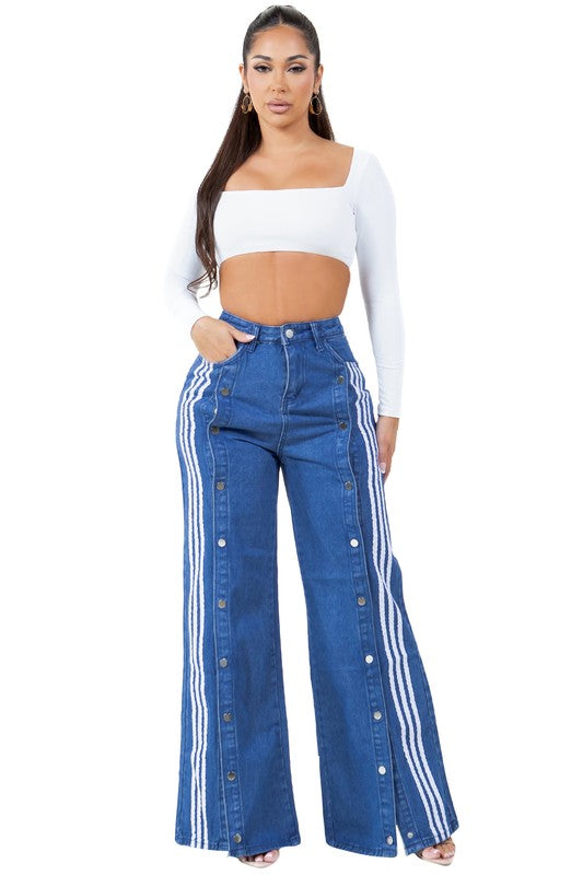 A woman with long dark hair wears a white cropped top paired with WOMEN FASHION CASUAL STYLE DENIM PANTS, which feature high-waist, wide-leg design with blue fabric adorned by white stripes and button details down the sides.