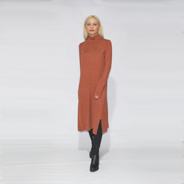 A person with long blonde hair is standing sideways, wearing a turtle neck dress with long sleeves and side slits in brown, paired with black boots, against a plain background.