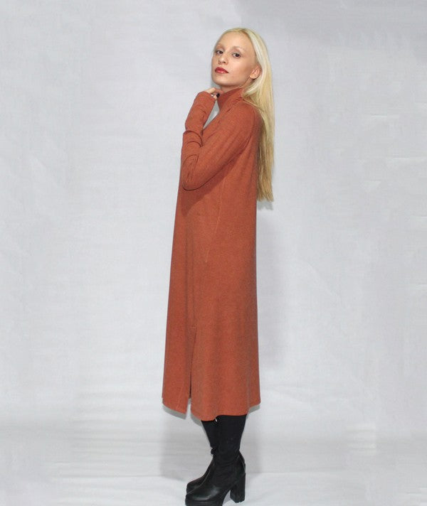 A person with long blonde hair is standing sideways, wearing a turtle neck dress with long sleeves and side slits in brown, paired with black boots, against a plain background.