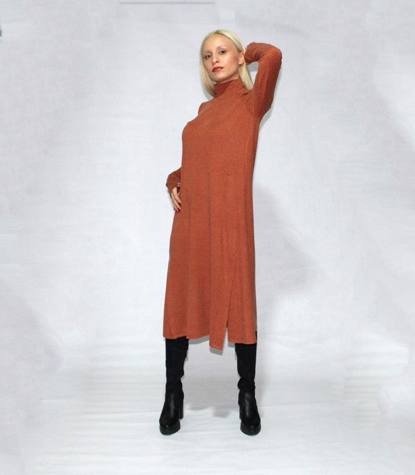 A person with long blonde hair is standing sideways, wearing a turtle neck dress with long sleeves and side slits in brown, paired with black boots, against a plain background.