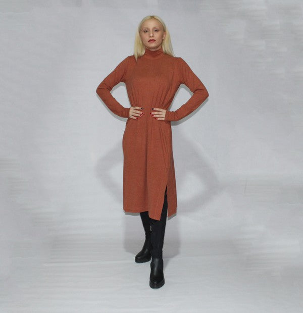 A person with long blonde hair is standing sideways, wearing a turtle neck dress with long sleeves and side slits in brown, paired with black boots, against a plain background.