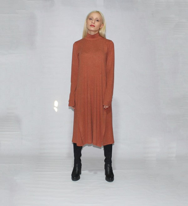 A person with long blonde hair is standing sideways, wearing a turtle neck dress with long sleeves and side slits in brown, paired with black boots, against a plain background.