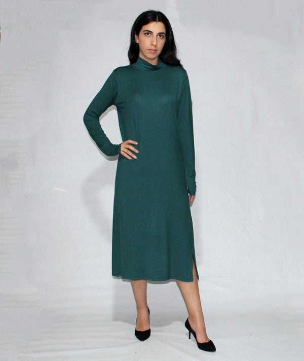 A woman in a Turtle neck dress long sleeve with side slits, teal and knee-length made from viscose, stands against a plain background, posing with one hand on her hip and wearing black heels.