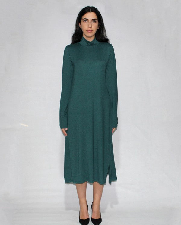 A woman in a Turtle neck dress long sleeve with side slits, teal and knee-length made from viscose, stands against a plain background, posing with one hand on her hip and wearing black heels.