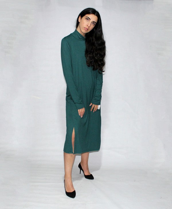 A woman in a Turtle neck dress long sleeve with side slits, teal and knee-length made from viscose, stands against a plain background, posing with one hand on her hip and wearing black heels.