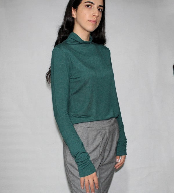 A person wearing gray high-waisted Extra SOFT dropped pants with press folds, black heels, and a green long-sleeve top stands with hands in pockets against a plain white background.